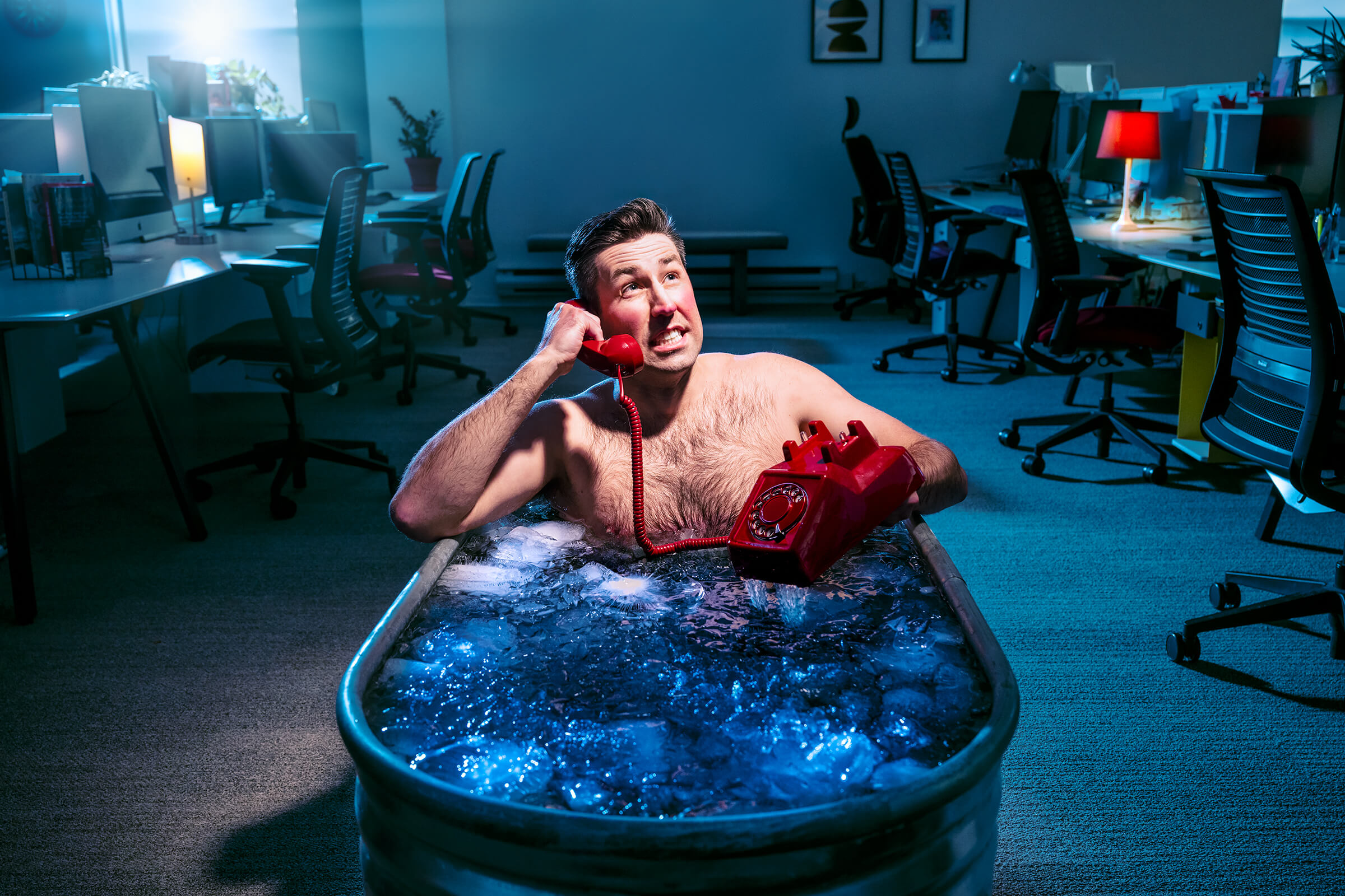 A guy in an icy bathtub in the middle of an office holding and talking on a rotary phone.