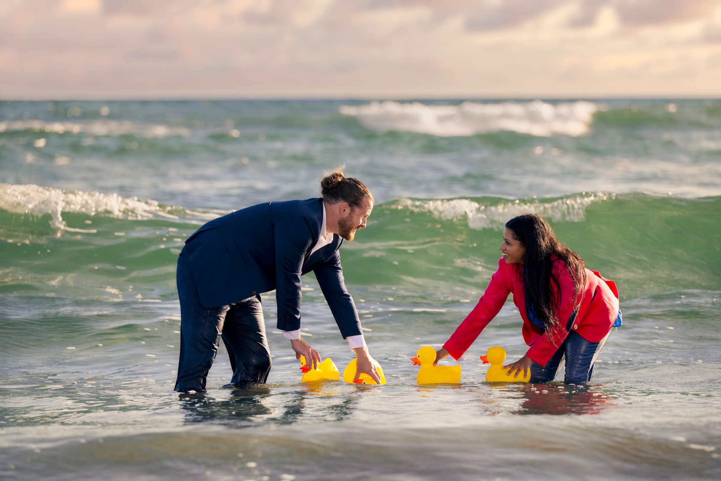 Two people on a beach in the waves holding four ducks that they're trying to place in a straight line.